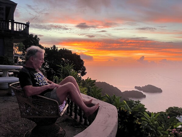 Un hombre sentado en un banco contemplando una puesta de sol con vistas al mar y a islas distantes.