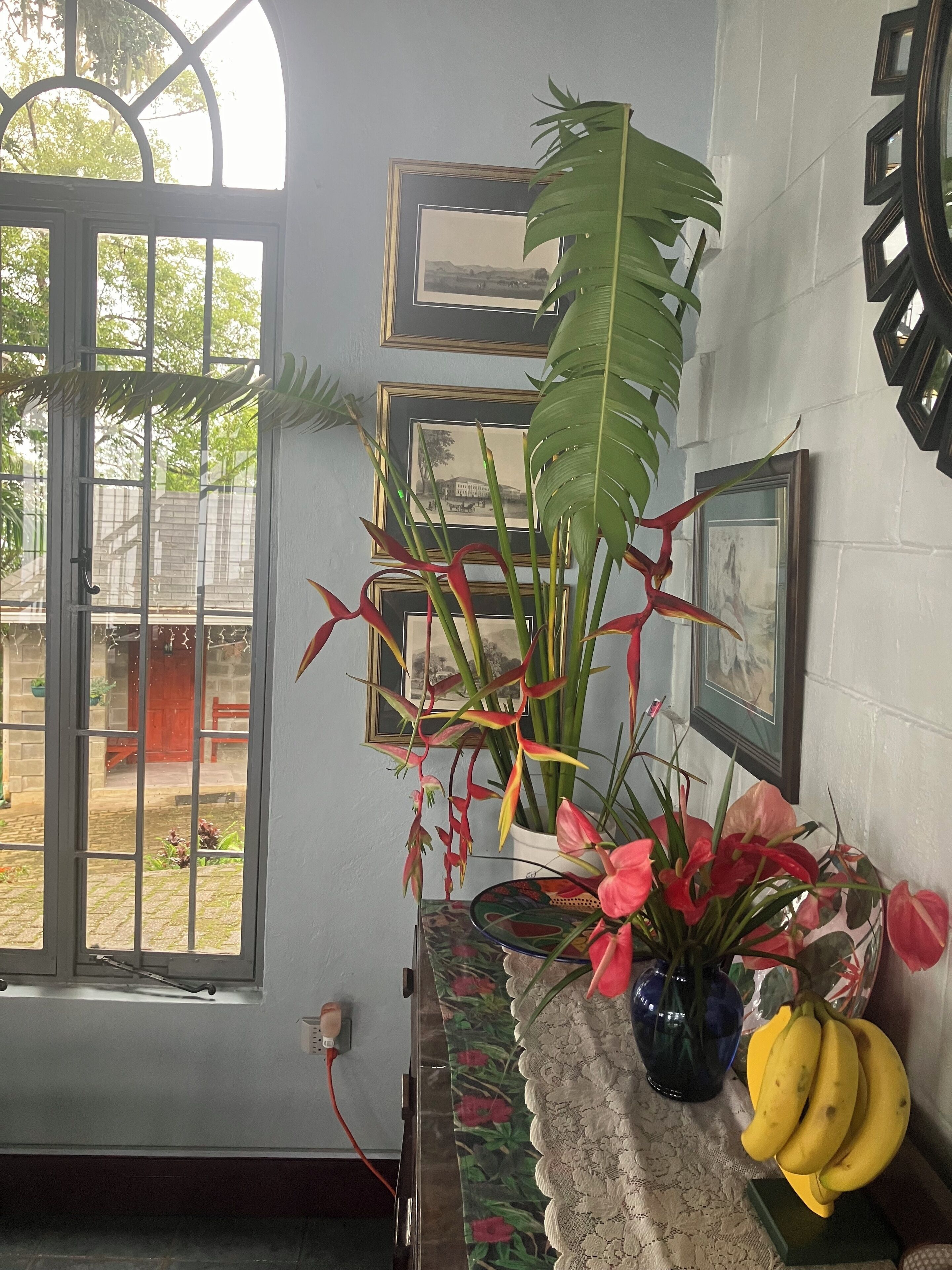 Una stanza con un'ampia finestra, una sveglia a muro e un tavolo con un vaso di fiori e banane.