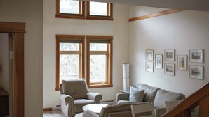 Ein Wohnzimmer mit einer Sofagarnitur, einem Couchtisch und einem Kamin. An den Wänden hängen Bilder und durch das Fenster ist ein Blick ins Grüne möglich.