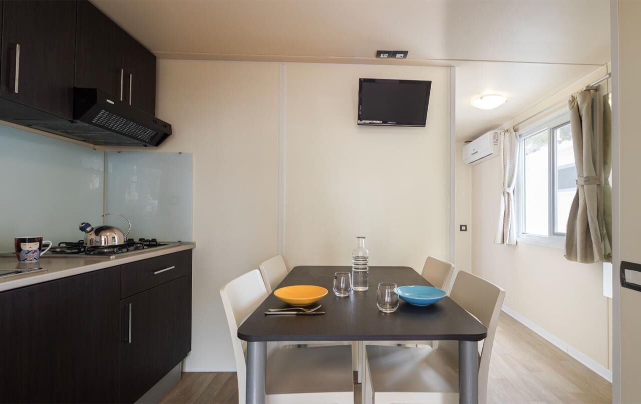 A compact kitchen with dark cabinets, a gas stove, a flat-screen TV, and a dining table with chairs.