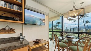 Habitación con televisión, una mesita de mimbre, una mesa de vidrio con sillas y vista a palmeras.