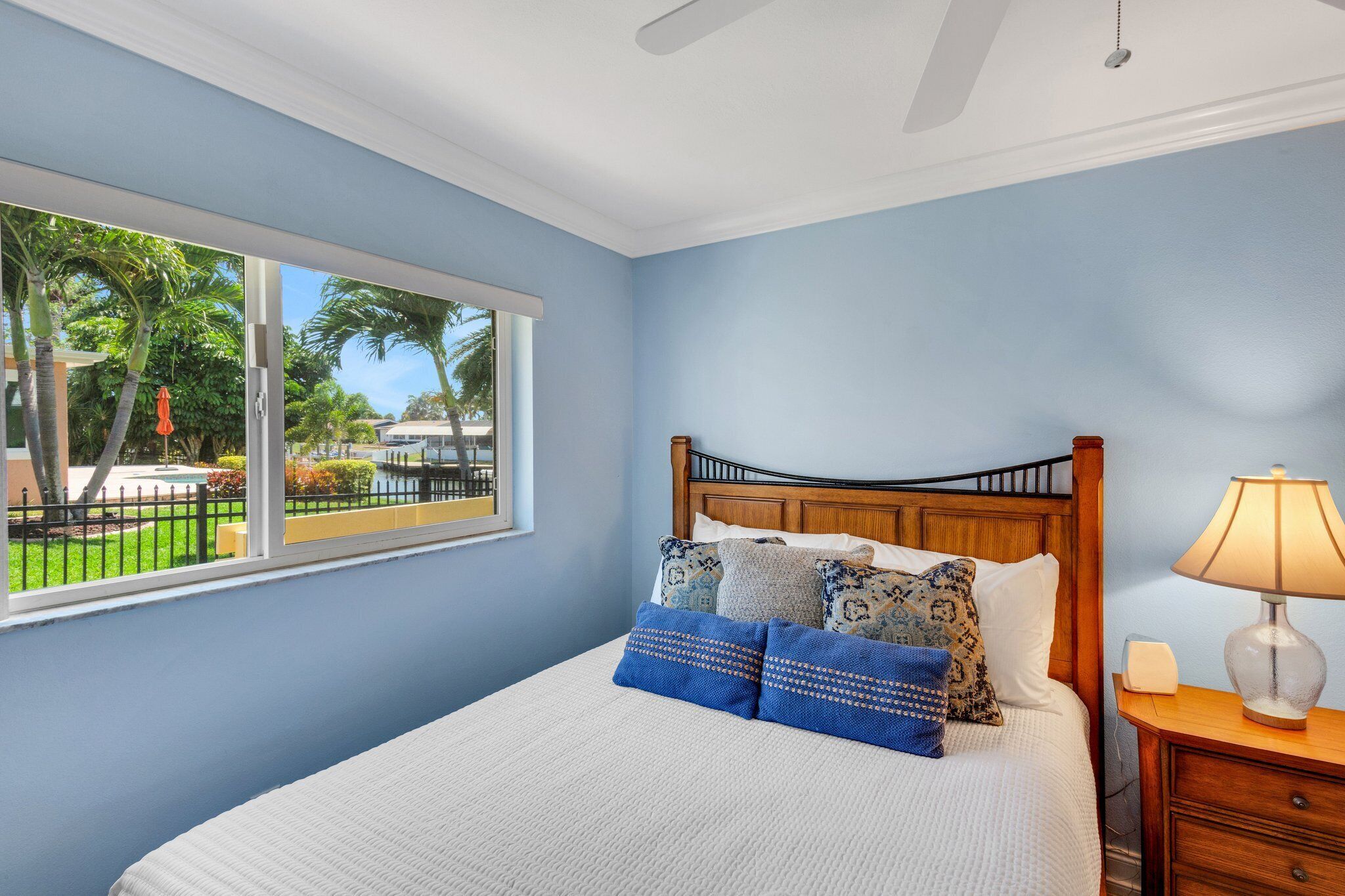 A bedroom with a bed, a nightstand, a lamp, and a window with a view of palm trees and a fence.