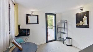 Een knus leefruimte met een eettafel, een klein keukenhoekje met een magnetron, een deur naar een buitenruimte, een metalen plank en een lijst met een foto aan de muur.