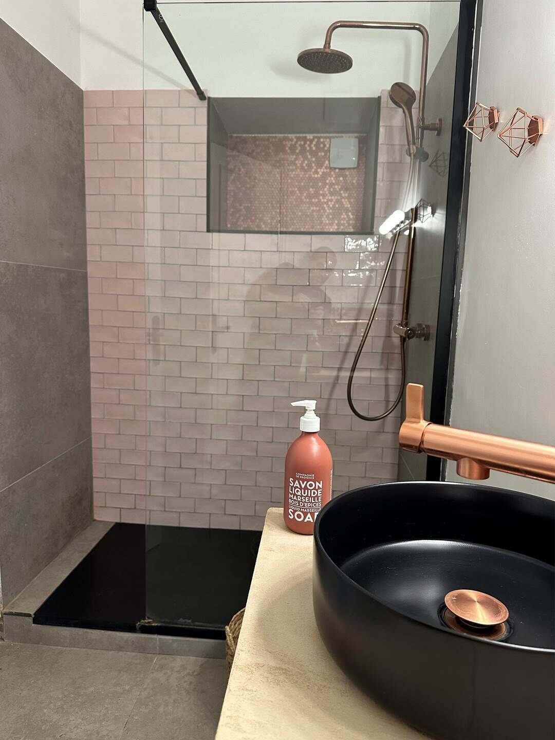 Ein modernes Badezimmer mit Duschkabine aus Glas, einem schwarzen Waschbecken mit Kupferarmatur und einer Seifenspende auf der Ablagefläche.