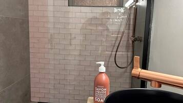 A modern bathroom with a glass shower enclosure, a black sink with a copper faucet, and a bottle of soap on the counter.