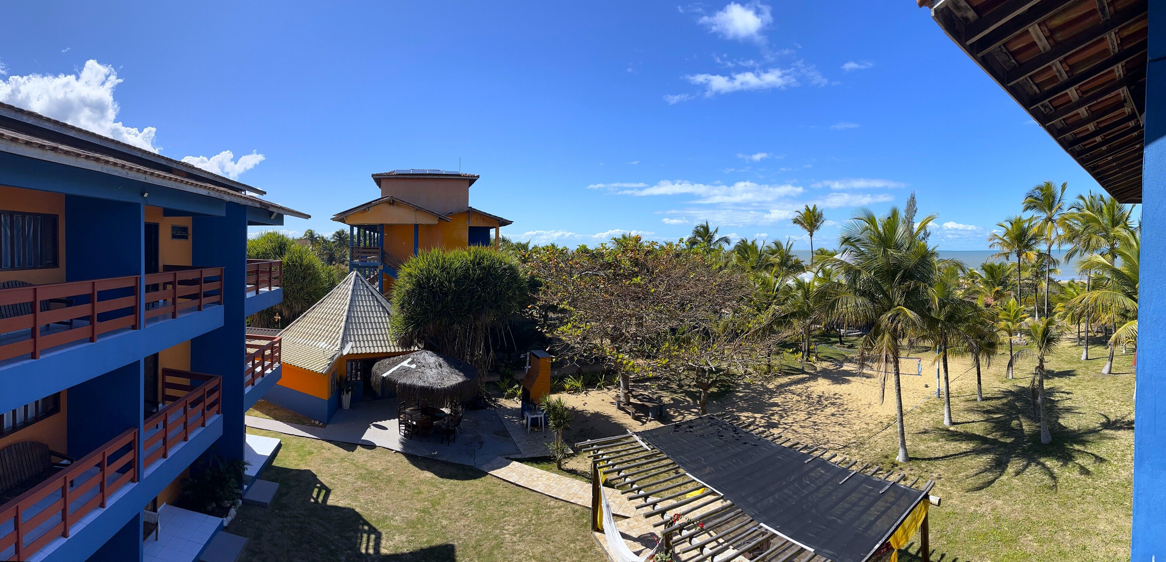 Um hotel com vários andares, área de piscina, construção com telhado de palha e vista para a praia com palmeiras.