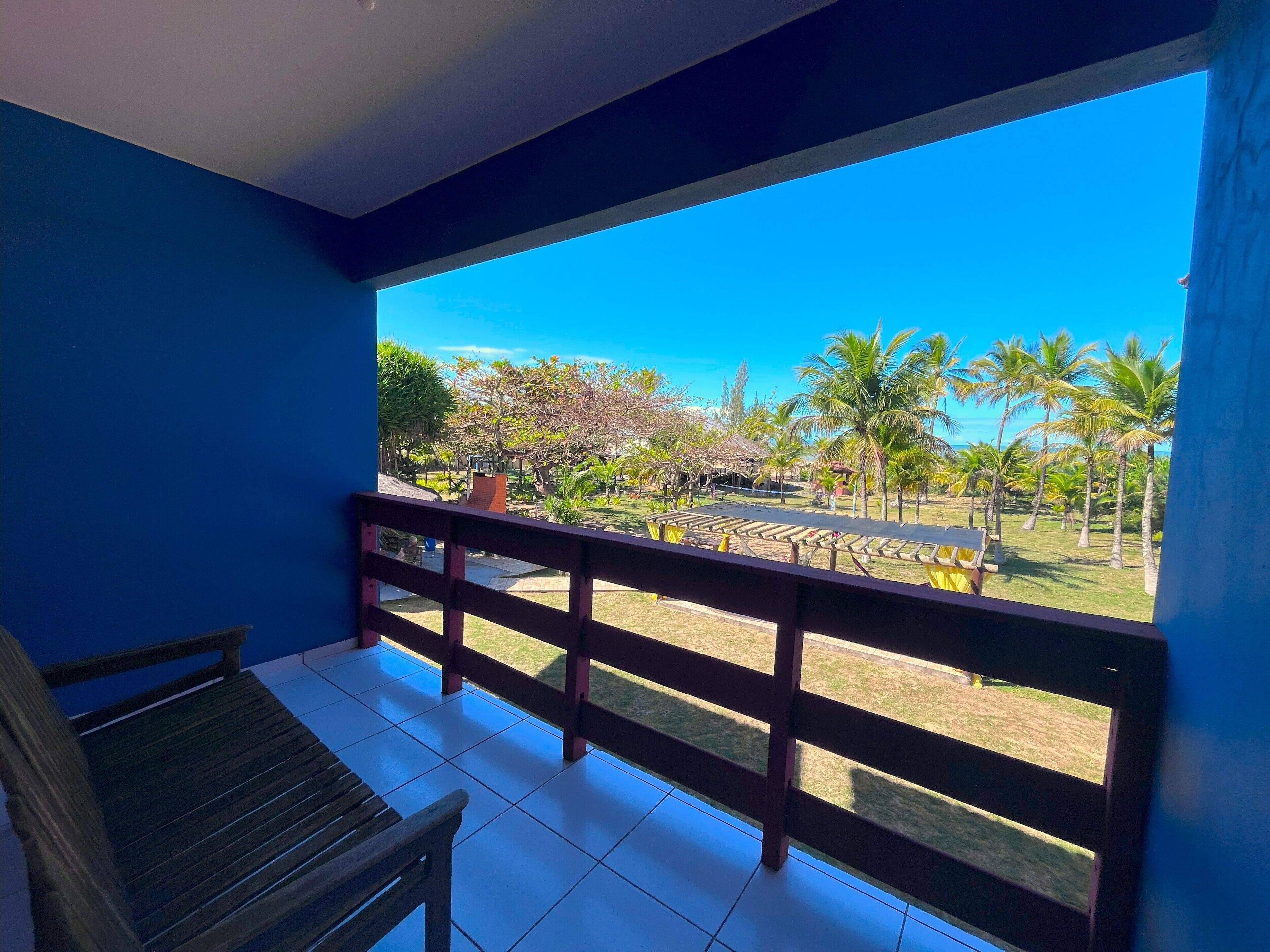 Een balkon met een houten bankje, een betegelde vloer en uitzicht op een tropische tuin met palmbomen en een helderblauwe lucht.
