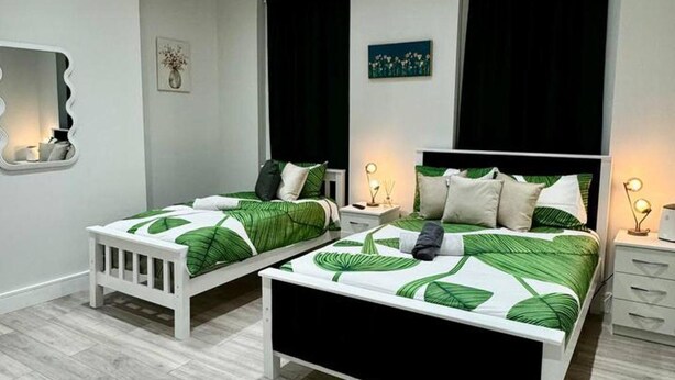 Une chambre à coucher avec deux lits simples, chacun doté d’un cadre blanc et d’une literie à motifs de feuilles vertes. On y trouve des tables de chevet avec des lampes, un petit tapis sur le sol et des œuvres d’art accrochées aux murs.