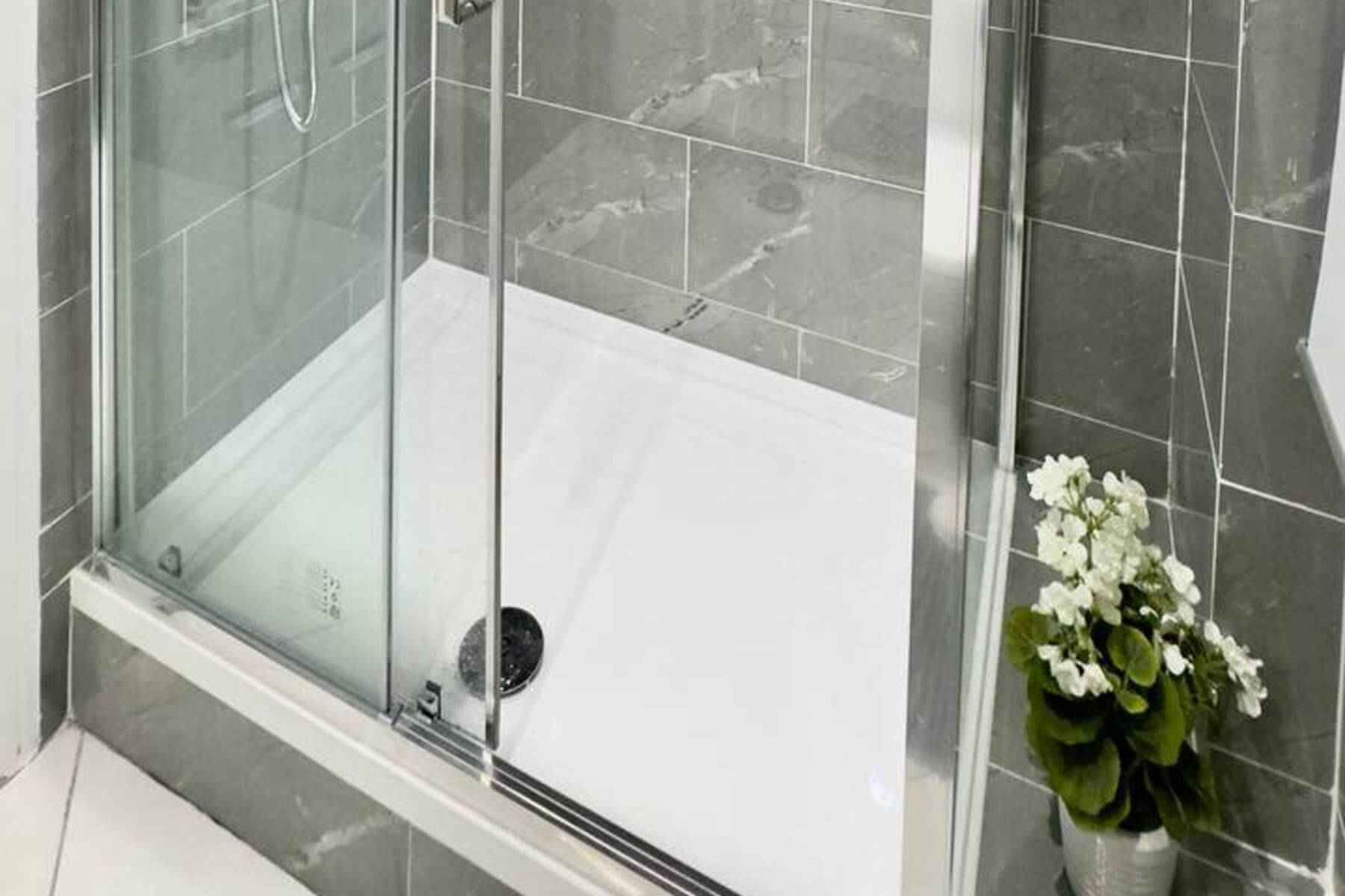 A glass-enclosed shower area with a white floor, grey tiles, and a potted plant with white flowers.
