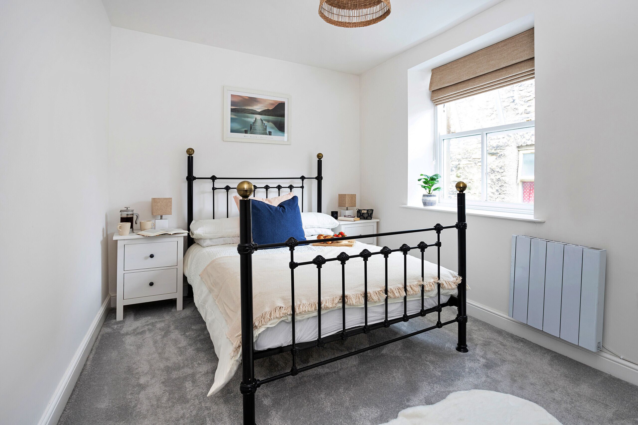 A bedroom with a black metal bed frame, white bedding, a bedside table with a lamp, and a window with a view of a stone wall.