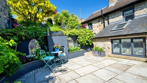 Une maison en pierre avec un patio, une petite table entourée de chaises et un jardin agrémenté de diverses plantes et fleurs.