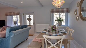 Un coin repas avec une table en bois dressée pour quatre personnes, entourée de chaises blanches. Un canapé bleu et un banc rose sont visibles au premier plan.