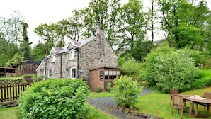 A stone house with a wooden door and windows, surrounded by lush greenery and a gravel path.