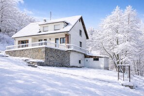 Uma casa de dois andares com base de pedra e uma varanda, rodeada por árvores cobertas de neve.