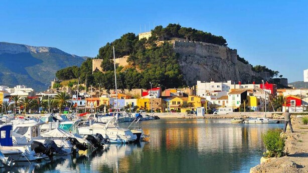 A marina with boats docked, a town with colorful buildings, and a hill with a fortification in the background. - Elegante Apartamento Frente al Puerto de Denia (Dénia)
