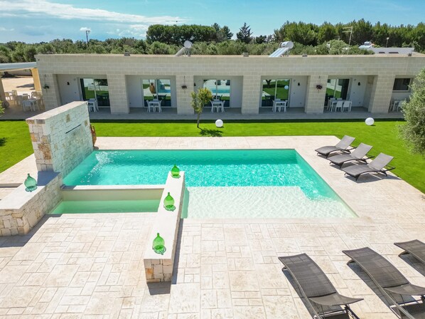 Une piscine rectangulaire bordée de pierres, entourée de chaises longues et d’un espace patio.
