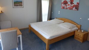 A bedroom with a wooden bed, a small bedside table, a chair, and a desk. There is a painting on the wall and a window with curtains.