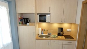 A compact kitchen with a sink, a microwave, a coffee maker, and a shelf with a kettle and a box of tissues.