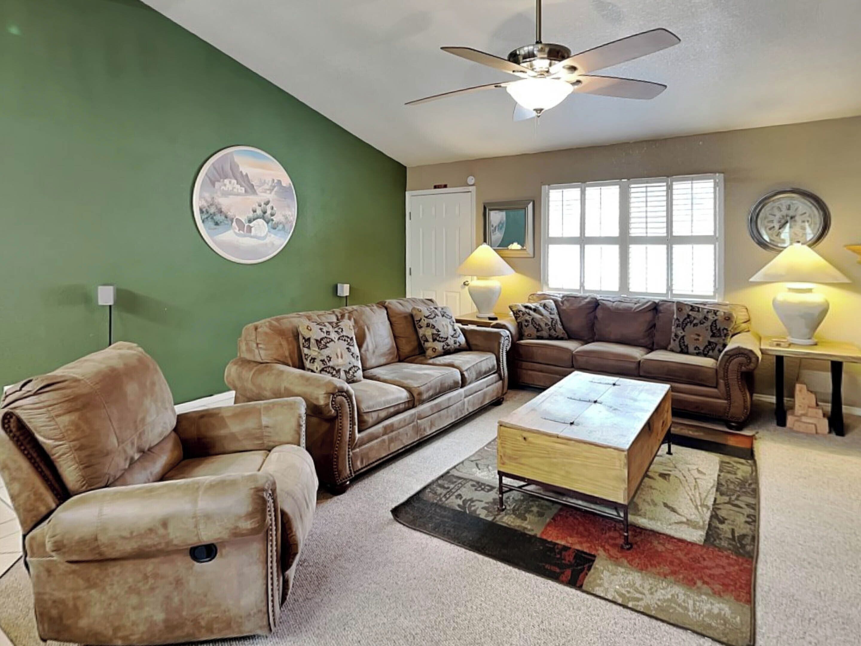 A living room with two sofas, a coffee table, a ceiling fan, a wall clock, and a window with blinds.