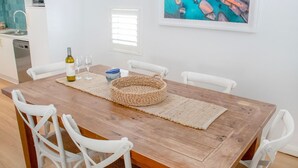 Un comedor con una mesa de madera, sillas blancas, un mantel tejido, una botella de vino y copas.