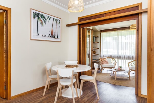A dining area with a round table and chairs, a framed picture on the wall, and a view into a living room with a sofa and a window.