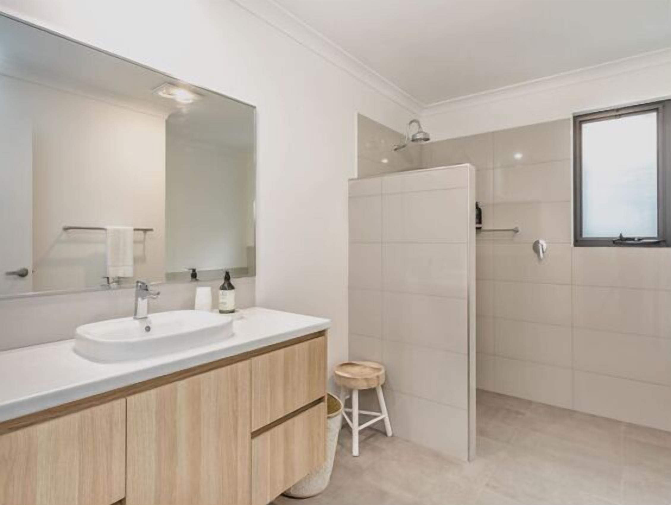 A modern bathroom with a large mirror, two sinks, a shower, and a small stool.