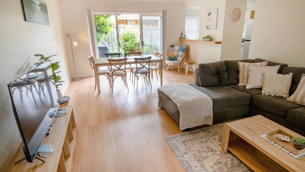A spacious living area with a wooden floor, a dark grey sofa, a coffee table, and a dining table with chairs. - This snug 3 bedroom villa is all you need to call home whilst visiting Albany (MIDDLETON BEACH)