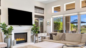 Een ruime woonkamer met een open haard, een flatscreen televisie en een groot raam met uitzicht op een tuin.