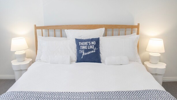 A neatly made bed with white bedding and a blue pillow with text, flanked by two bedside tables with lamps, in a room with a simple, clean design.