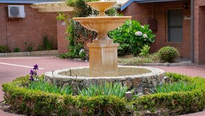 A fountain with a circular stone base surrounded by greenery, set against a brick building with a blue roof.