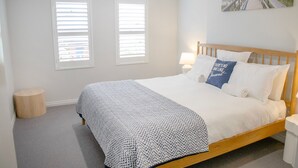 A bedroom with a wooden bed, white bedding, two windows with blinds, a round wooden stool, and a beach-themed painting on the wall.