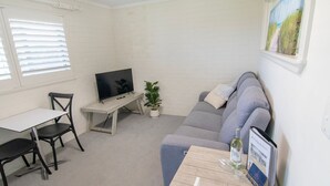 Ein Zimmer mit grauem Sofa, einem kleinen Tisch mit Flasche und Gläsern, einem Fernseher auf einem Holzboden, einer Zimmerpflanze und einem Fenster mit Jalousien.