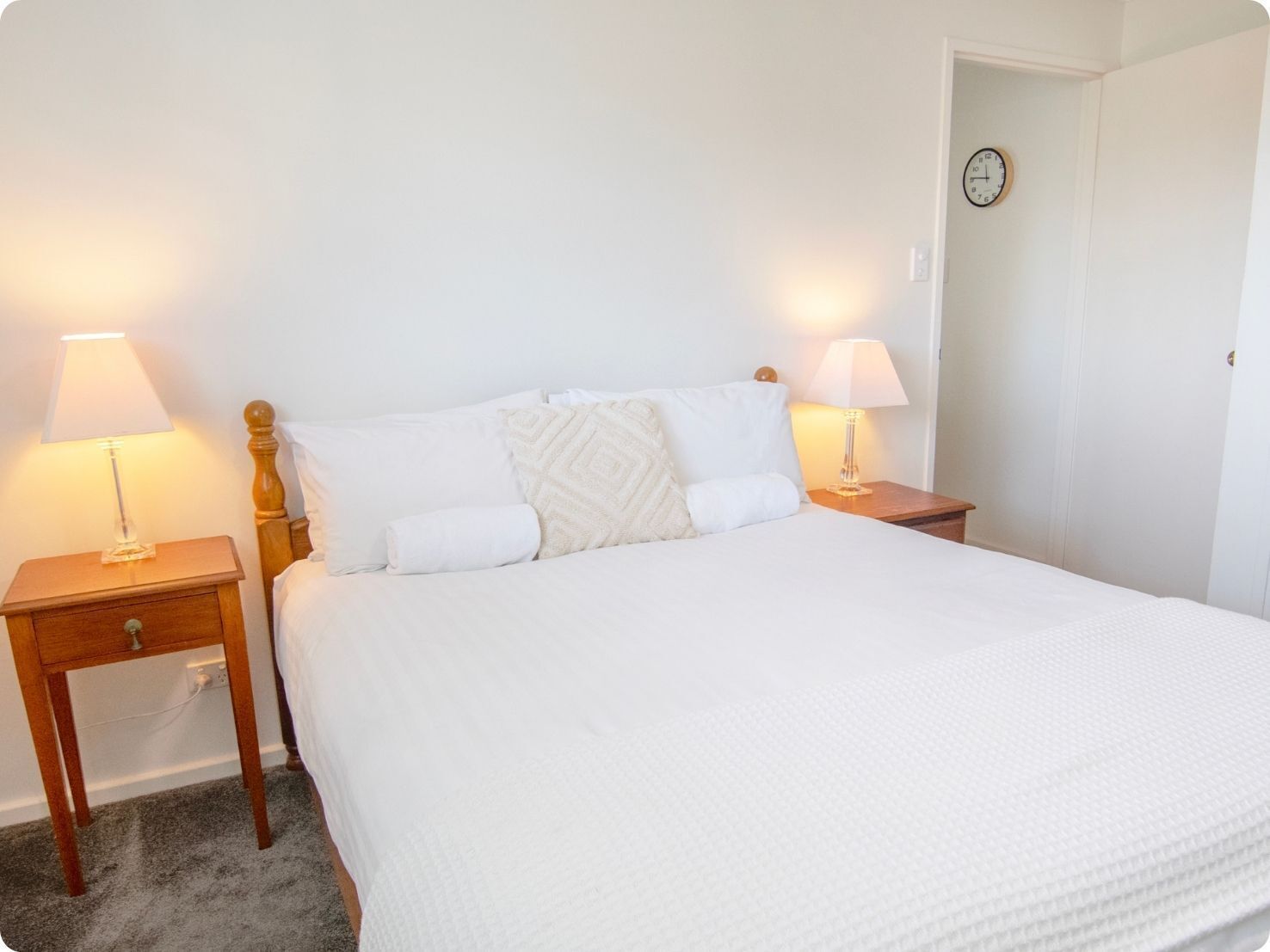 A neatly made bed with white bedding, two bedside tables with lamps, and a clock on the wall.
