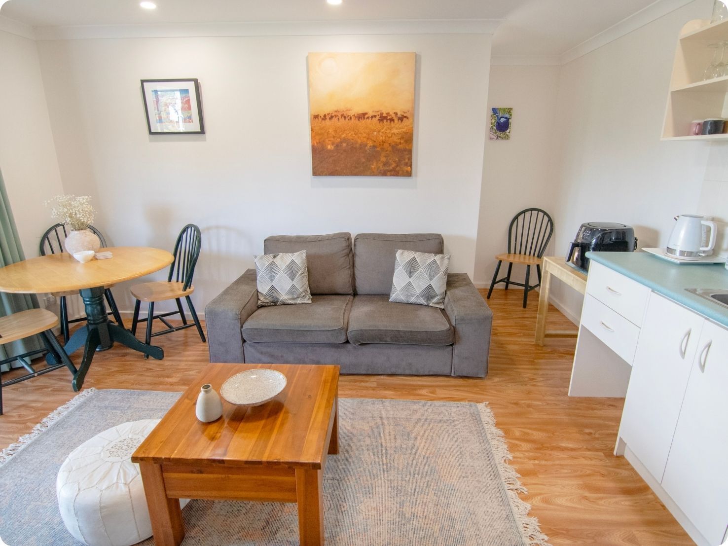 A cozy living area with a grey sofa, a wooden coffee table, and a dining table with chairs. A painting hangs on the wall, and a kitchen area is visible in the background.