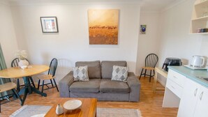 A cozy living area with a grey sofa, a wooden coffee table, and a dining table with chairs. A painting hangs on the wall, and a kitchen area is visible in the background. - Cute upstairs 2 bedroom apartment perfect for short stays while visiting Albany (MIRA MAR)