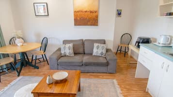 A cozy living area with a grey sofa, a wooden coffee table, and a dining table with chairs. A painting hangs on the wall, and a kitchen area is visible in the background.