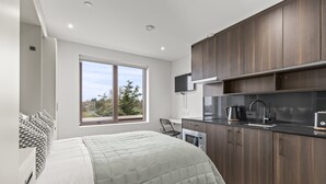 A modern bedroom with a large bed, a window with a view, and a kitchenette with dark wood cabinets and a black countertop. - Executive Apartment (London)