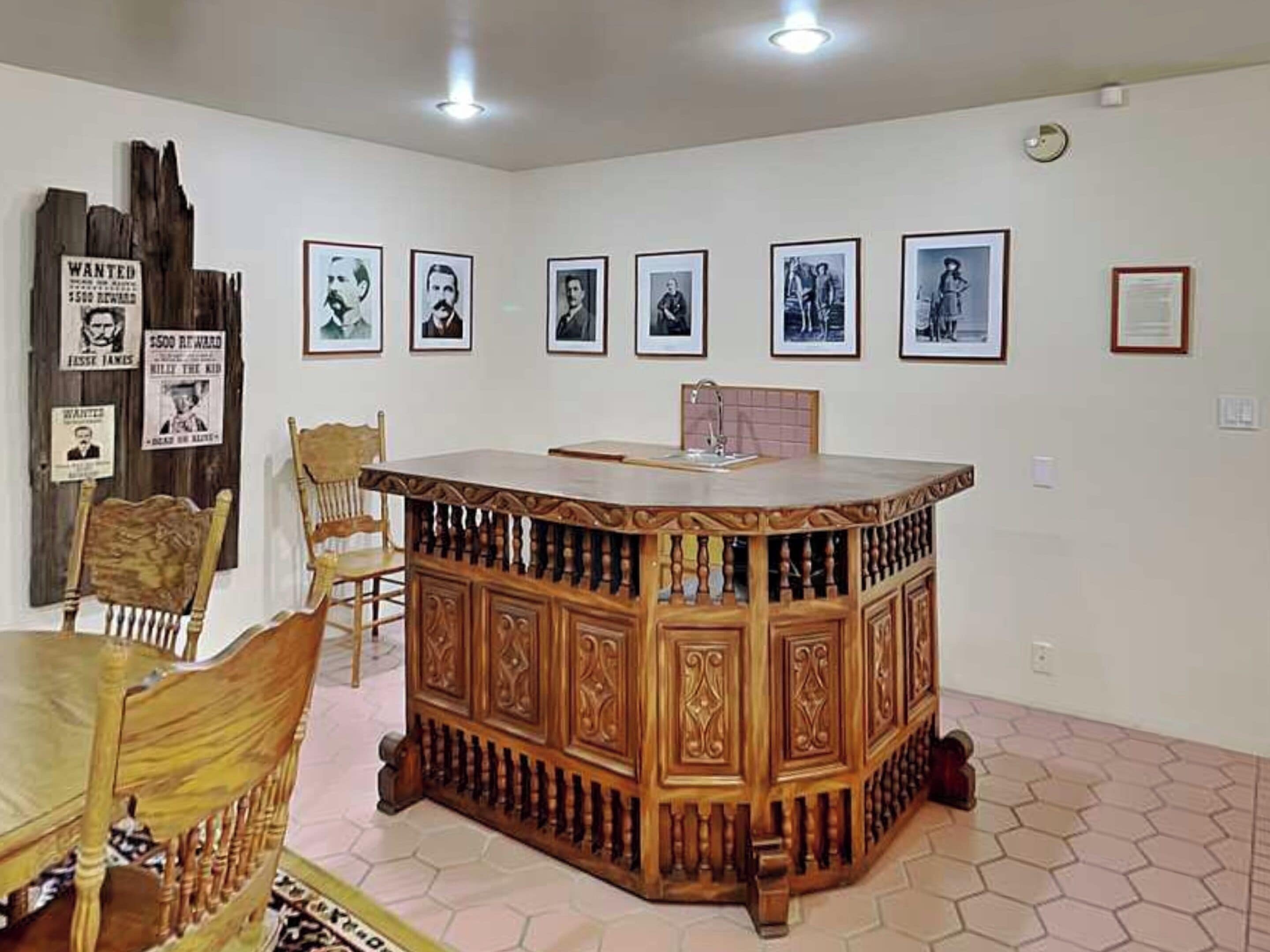 A room with a wooden bar, a dining table, chairs, and a hexagonal patterned floor. There are framed pictures on the wall.
