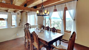 Comedor con mesa y sillas de madera, una araña y ventanas amplias con cortinas.