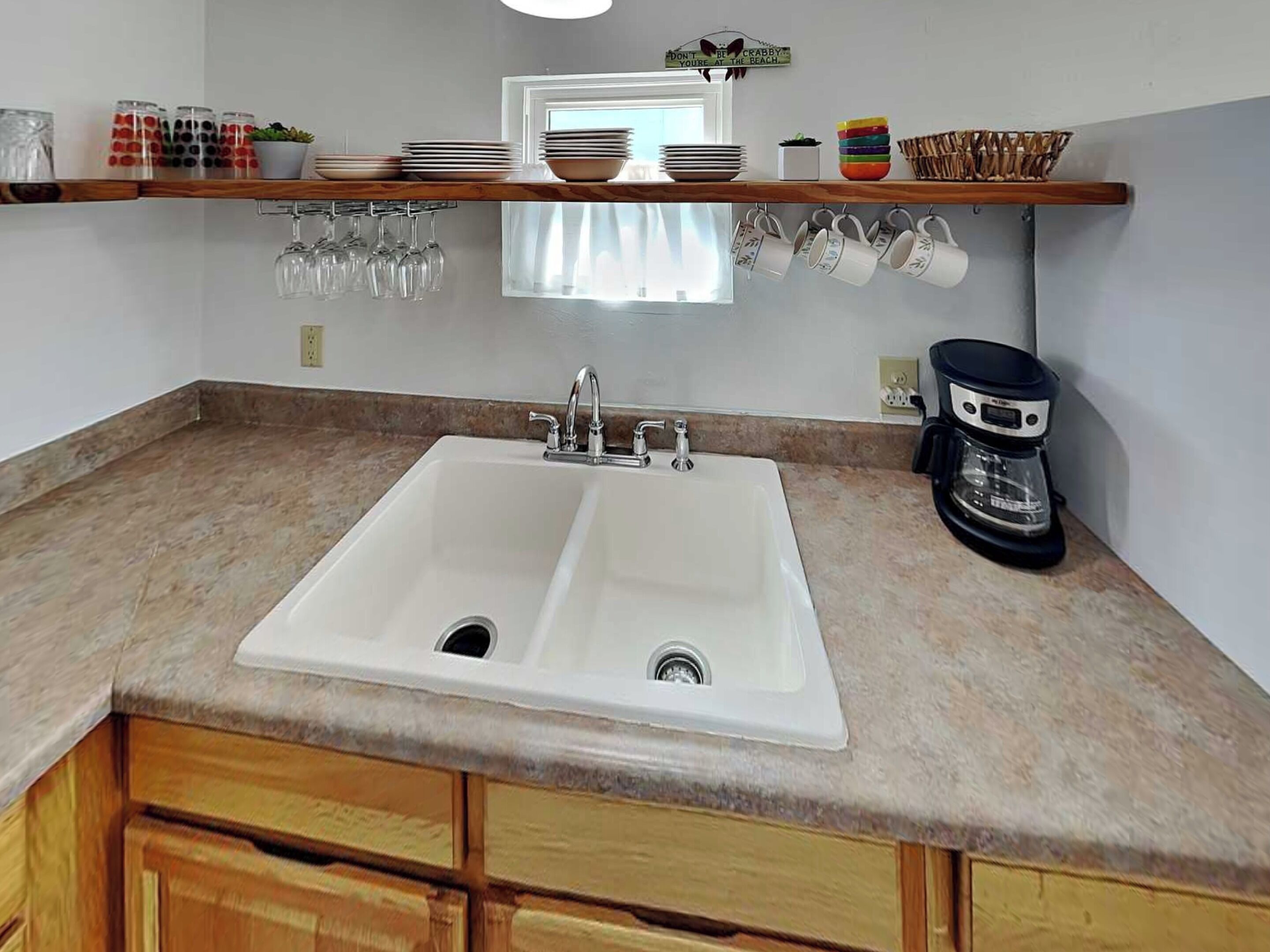 A kitchen with a double sink, wooden cabinets, a coffee maker, and a shelf with dishes and glasses.