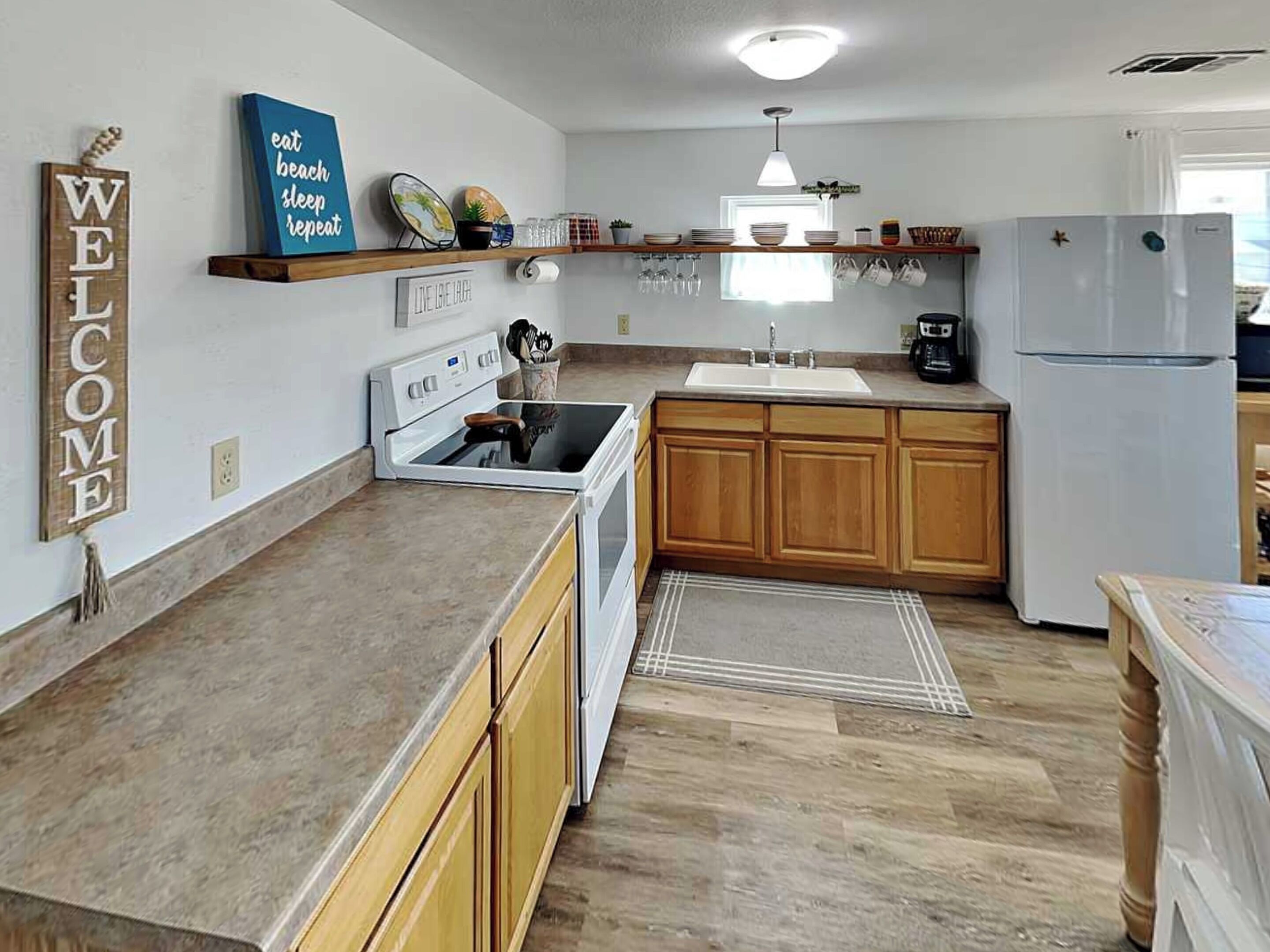 Una cocina moderna con armarios de madera, horno empotrado, pileta y refrigerador. Hay elementos decorativos en las repisas y un cartel de bienvenida en la pared.
