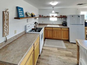 A modern kitchen with wooden cabinets, a built-in oven, a sink, and a refrigerator. There are decorative items on the shelves and a welcome sign on the wall.