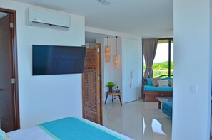 A hotel room with a bed, a television, an air conditioning unit, a small table with a plant, and a sofa with a view of greenery outside. - Heart designed Apartment, Ocean and Lagoon view, large terrace  (Cancún)