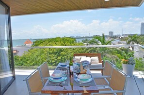 A balcony with a dining table set for two, overlooking a cityscape and a body of water. - Heart designed Apartment, Ocean and Lagoon view, large terrace  (Cancún)