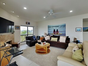 Un salon moderne avec une cheminée, une grande fenêtre, un ventilateur de plafond et un tableau sur le thème de la plage.