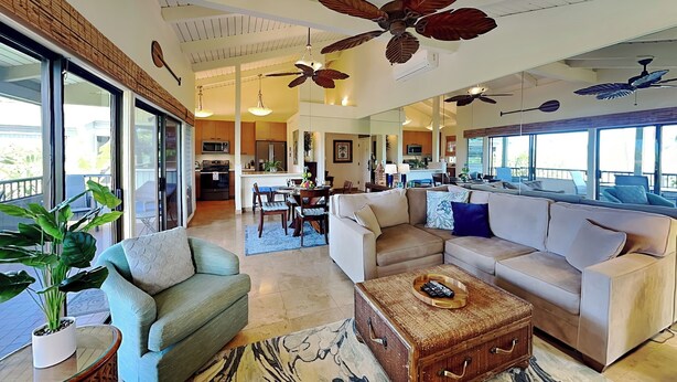 A spacious living area with a large sofa, a coffee table, and a dining table. There are ceiling fans, a kitchen in the background, and large windows. - Wailea Ekolu Village 1406 by Maui Rental Group (Kihei)