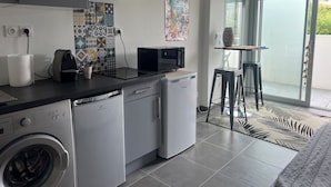 Eine kompakte Küche mit Waschmaschine, Mikrowelle und Kaffeemaschine. Ein kleiner Tisch mit einer Vase und einem Stuhl stehen bereit. Das Zimmer verfügt über ein großes Fenster und einen gemusterten Teppich.