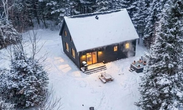 A cabin with a snow-covered roof, a porch with a fire pit, and a hot tub.