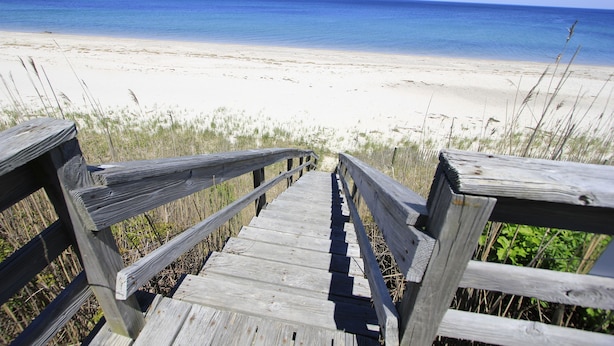 Ein Holzsteg führt zu einem Strand mit klarem, blauem Wasser.