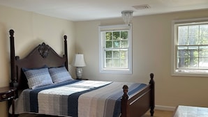 A bedroom with a wooden bed, a nightstand, a lamp, and a window with a view of trees. - Beautiful sunset on John’s Pond (Mashpee)